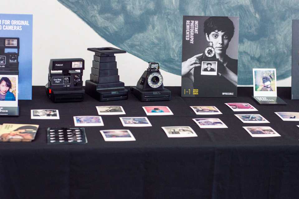 Books, instant film photographs, a Polaroid One Step camera, an Impossible Instant Lab camera and an Impossible I-1 camera line a table at PolaCon. 