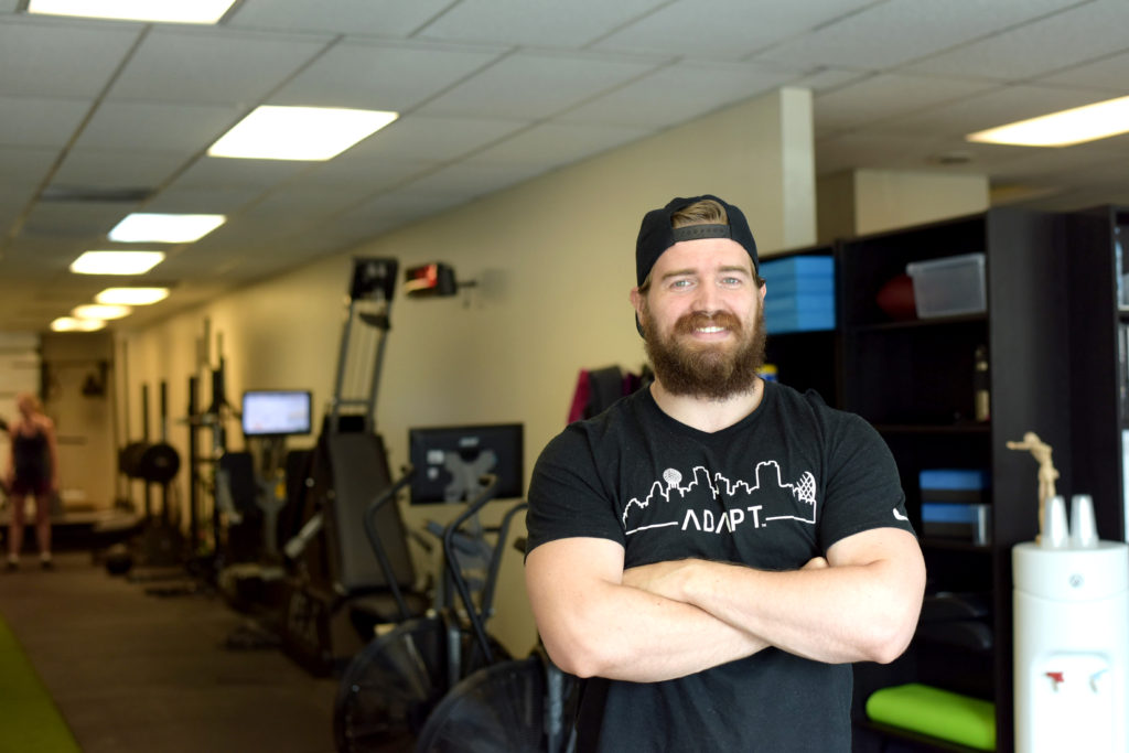 Alexander, a burly man with a red mustache and beard, stands smiling with his arms crossed inside his gym, with assorted physical training equipment in the background.