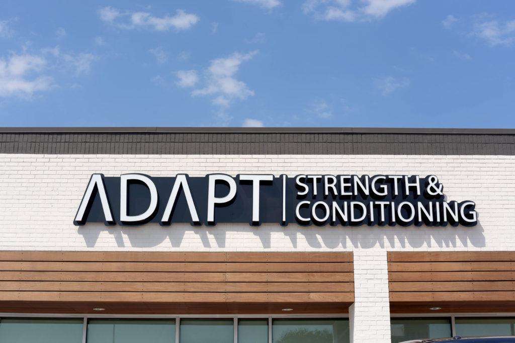 ADAPT Strength & Conditioning, printed in a white, modern font above wooden awnings on turquoise windows.