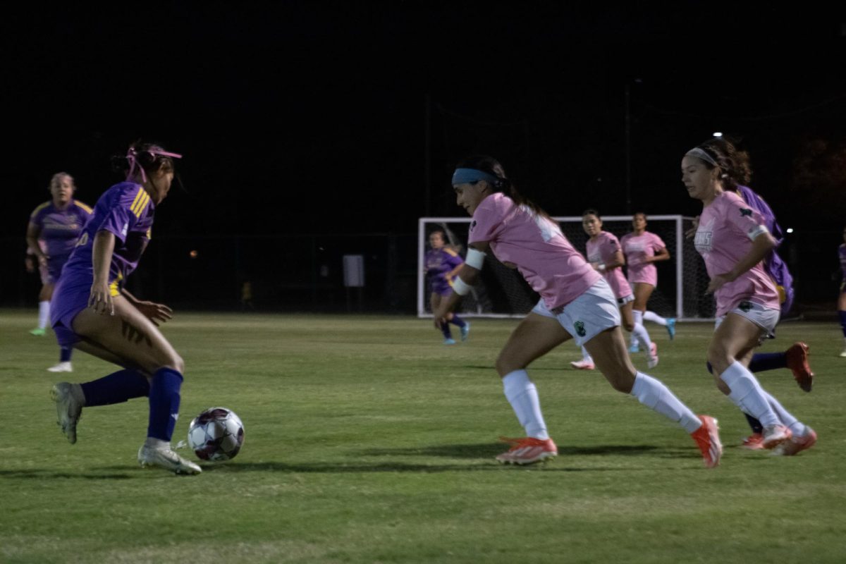 A girl in violet uniform kicking the ball and a girl in pink uniform trying to block the ball.